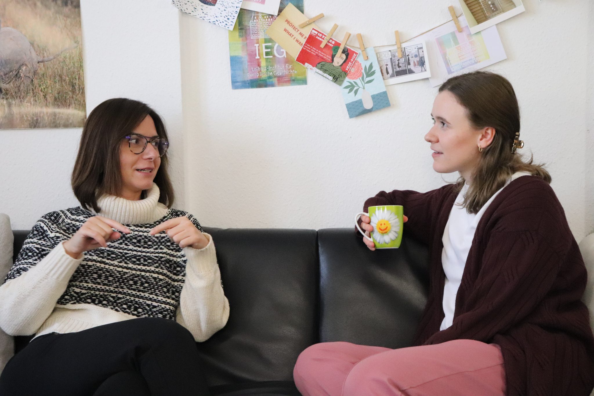 zwei junge Frauen sitzen auf einem Sofa und unterhalten sich. Im Hintergrund hängen Postkarten und Bilder. Eine der Frauen hat eine Kaffeetasse in der Hand.