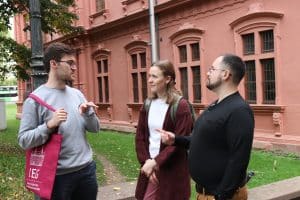 Drei Stipendiaten stehen draußen und unterhalten sich. Zwei Männer und eine Frau. Einer der Männer trägt einen roten IEG Jutebeteutel.