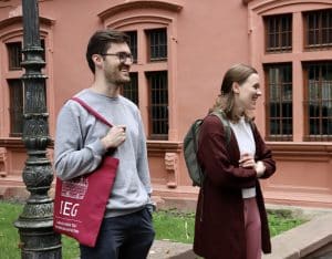 ein junger männlicher Stipendiat mit einem roten IEG Beutel steht vor einem historischen Gebäude und lacht. Neben ihm steht eine junge Frau mit Rucksack und blickt ebenfalls lachend zur Seite zu einem Gesprächspartner außerhalb des Bildes.